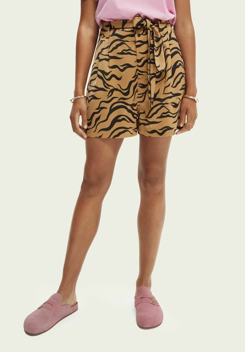 Femme portant un short imprimé tigre marron avec une ceinture assortie, un haut rose, des bracelets dorés et des chaussures roses sans lacets.