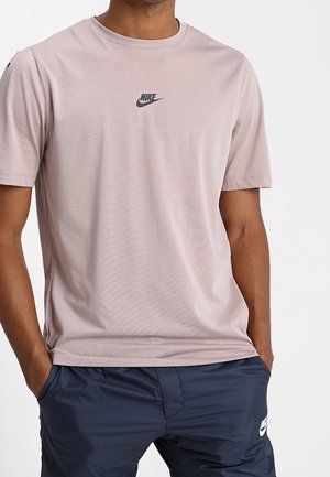 Man draagt een lichtroze Nike T-shirt en donkerblauwe Nike short met handen in de zakken, staand tegen een effen achtergrond.