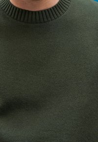 Gros plan sur une personne portant un pull tricoté vert foncé côtelé avec un col rond.