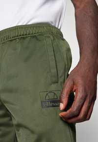 Olijfgroene shorts met een elastische tailleband en zijzakken. Bevat een zwart logo op de rechter onderzijde van het been. Het materiaal lijkt lichtgewicht te zijn.