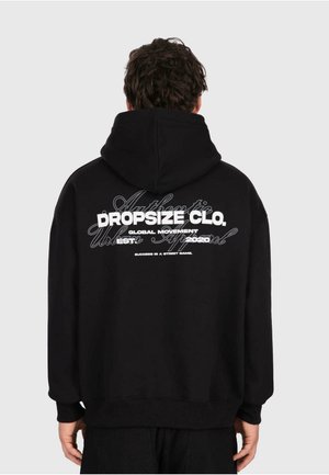 Persona che indossa un hoodies nero con testo bianco sulla schiena: "Dropsize Clo. Movimento Globale Fondato nel 2020" e un testo corsivo più piccolo.