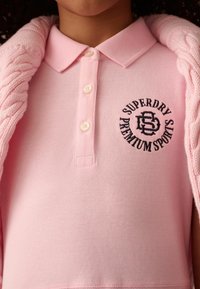 Polo tričko svetloružovej farby z bavlny s krátkymi rukávmi, s dvoma gombíkmi a okrúhlym vyšitým logom tmavšej ružovej.