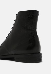 Botín negro de cuero hasta el tobillo con una superficie texturizada, acentos suaves, puntera redonda y un bajo tacón de madera. Diseño de cordones en la parte delantera.