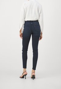 Jeans skinny de cintura alta en denim de lavado oscuro, con bolsillos traseros y una textura suave, combinados con una blusa blanca de manga larga y tacones.