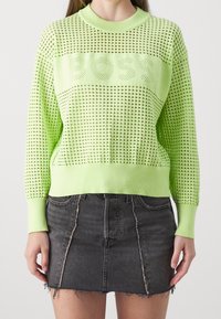 Maglione a maglia verde lime con design a rete aperta e logo BOSS, abbinato a una gonna di jeans nera con orlo sfrangiato e cuciture frontali.