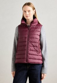 Naine kannab käeaukudega marooni värvi tikitud vestit, mille all on hallast kudumistehtud sokk ja tume sinine teksadega, lihtsa heleda tausta ees.