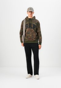 Fiatal férfi terepmintás GAP kapucnis pulóverben, fekete nadrágban, fehér slip-on cipőben, szürke sapkában, és szürke válltáskával a sima fehér háttér előtt.