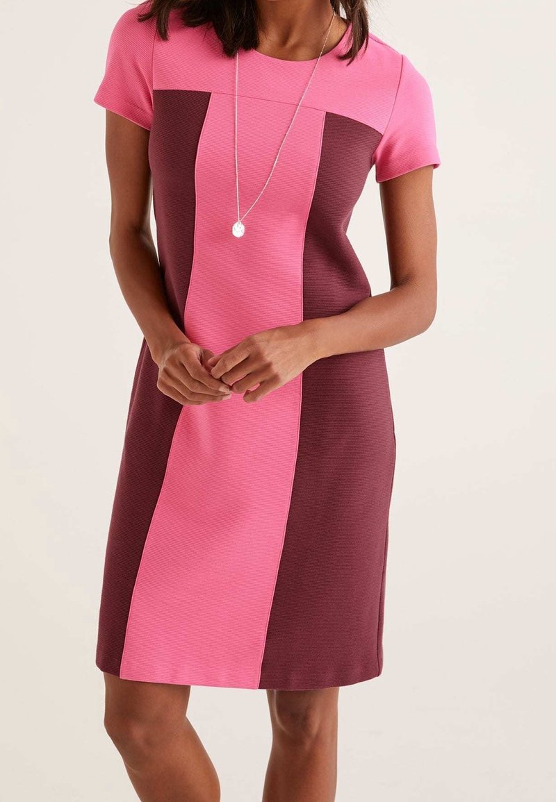 Femme portant une robe courte à manches courtes, longueur genou, avec des blocs de couleurs verticaux rose et bordeaux et un collier pendentif.