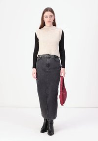 Maglione senza maniche beige chiaro sopra una maglietta nera a maniche lunghe, abbinato a una gonna in denim scuro e stivali neri alla caviglia, con una borsa rossa in mano.