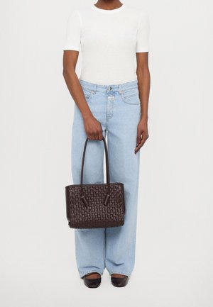 Personne portant un haut blanc à manches courtes, un jean large bleu clair, des ballerines noires, tenant un sac fourre-tout tissé marron foncé par les poignées.