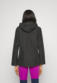 Giacca impermeabile nera con cappuccio, caratterizzata da una texture liscia e un design aderente. Abbinata a leggings vibranti viola e blu.