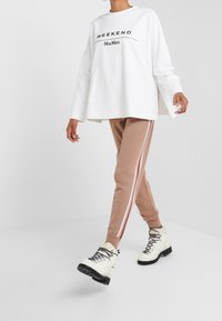 Sudadera blanca con el texto "WEEKEND" y logo, combinada con joggers beiges que tienen rayas laterales blancas y rojas, y botas blancas con cordones.