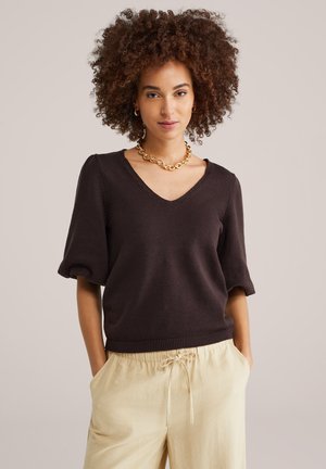 Žena s kučeravými vlasmi v tmavom svetri s výstrihom do V, béžových nohaviciach so sťahovacou šnúrkou, zlatom reťazovom náhrdelníku a malých kruhových náušniciach, ruky v kapsách.