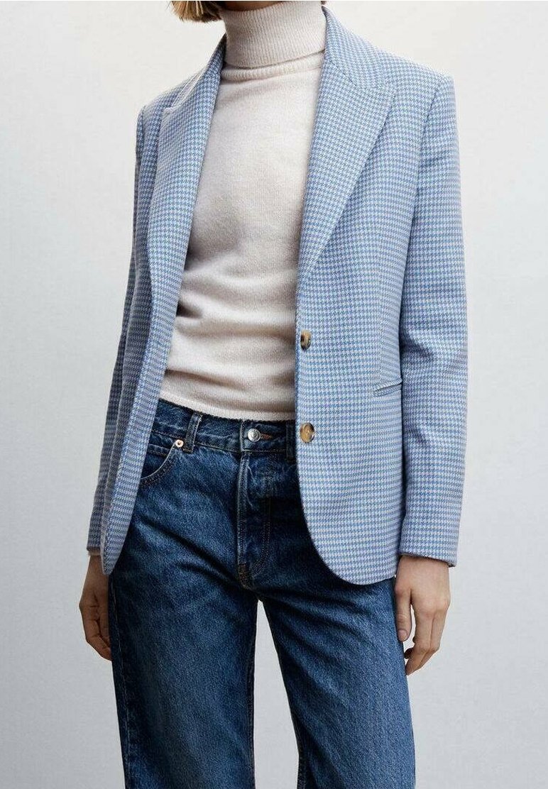 Femme portant un blazer pied-de-poule bleu clair sur un pull à col roulé beige et un jean bleu taille haute, debout devant un fond uni.