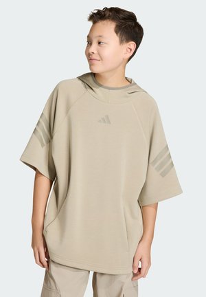 Niño con sudadera beige de manga corta y capucha de gran tamaño con el logo de Adidas, mirando hacia un lado, de pie contra un fondo claro.