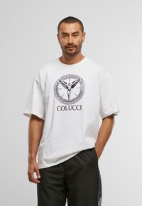 Vīrietis ar īsiem matiem un bārdu, kas ģērbts baltā Colucci t-kreklā ar spārnotā figūras logotipu, melnās biksēs, roka kabatā pret vienkāršu baltu fonu.