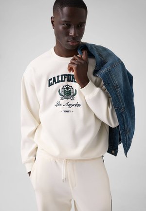 Jeune homme portant un sweat-shirt et un pantalon beige clair California Los Angeles, tenant une veste en jean bleue sur son épaule devant un fond uni.