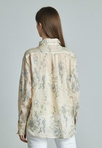 Blusa beige chiaro con ricami floreali in blu e verde. Presenta un colletto, polsini abbottonati e una vestibilità rilassata. Materiale in lino testurizzato.