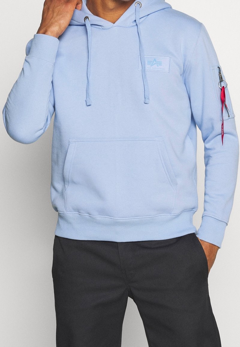 Felpa azzurra chiara con tasca frontale, cordini e tasca con zip sul braccio. Realizzata in tessuto morbido, con dettagli del logo sul petto.