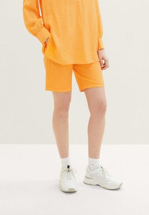 Pomarańczowe, lekkie szorty połączone z dopasowaną pomarańczową, oversize'ową koszulą i białymi trampkami z szarymi akcentami. Gładka tekstura, casualowy design.