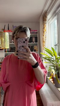 Rožinė, laisva suknelė su trumpomis, pūkuotomis rankovėmis ir laisvu siluetu. Lygi medžiaga švelniai krenta. Ilgi plaukai ir raštuotas telefonų dėklas rankoje.