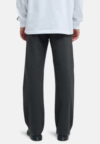 Persoon met de rug naar de kijker, gekleed in losse zwarte broek, zwarte schoenen en een wit shirt met lange mouwen, handen ontspannen langs het lichaam.