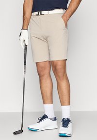 Μπέιζ κοντοπαντελονάκια γκολφ με τσέπες, συνδυασμένα με λευκή ζώνη και λευκά παπούτσια με μπλε λεπτομέρειες. Χέρι με γάντι που κρατάει ένα μπαστούνι γκολφ.