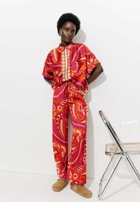 Ensemble de chemise et pantalon à motifs rouges et roses, présentant des designs floraux et paisley, associé à des chaussures pelucheuses marron. Détails de boutons simples.