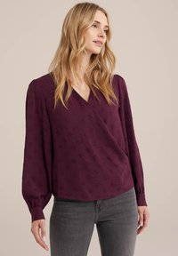 Burgunderfarbene Bluse mit Wickeldesign, langen Puffärmeln, strukturierter Stoff und feinem Muster. Kombiniert mit grauen Jeans, die eine entspannte Passform betonen.