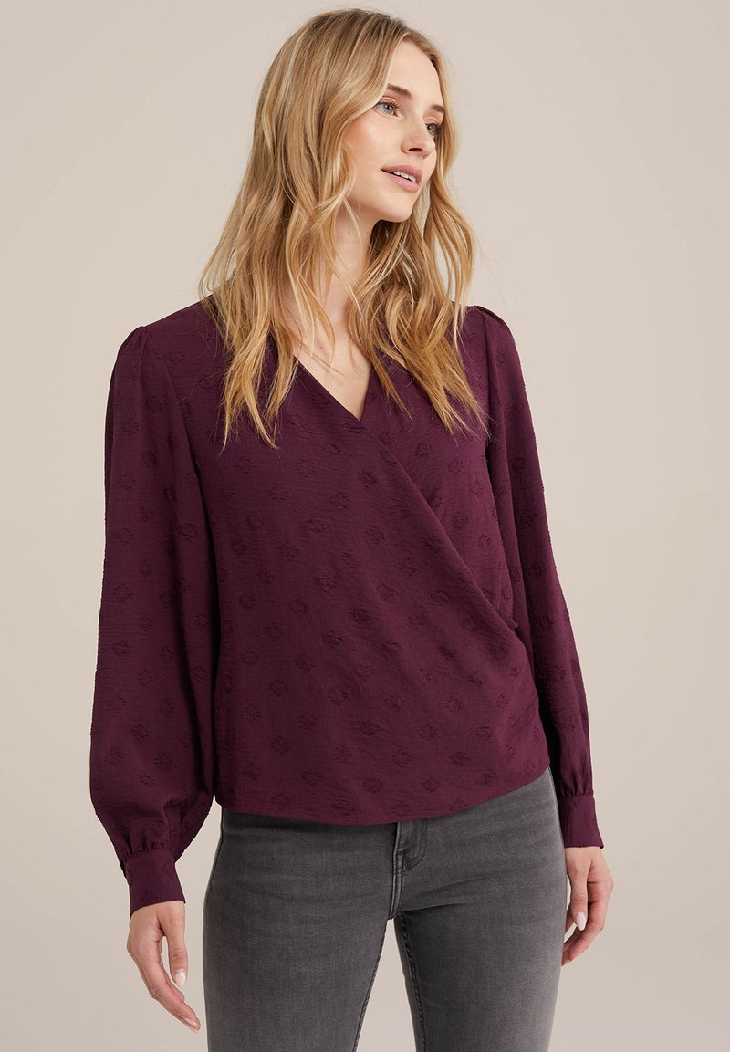 Burgunderfarbene Bluse mit Wickeldesign, langen Puffärmeln, strukturierter Stoff und feinem Muster. Kombiniert mit grauen Jeans, die eine entspannte Passform betonen.