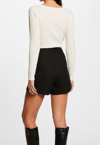 Mujer con una camiseta de manga larga blanca de canalé, pantalones cortos negros de alta cintura con textura y botas negras hasta la mitad de la pantorrilla, vista desde atrás.