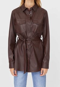 Brązowa skórzana koszula-kurtka z kołnierzykiem, zapinana na guziki z dwoma kieszeniami na piersi i paskiem wiązanym w talii.