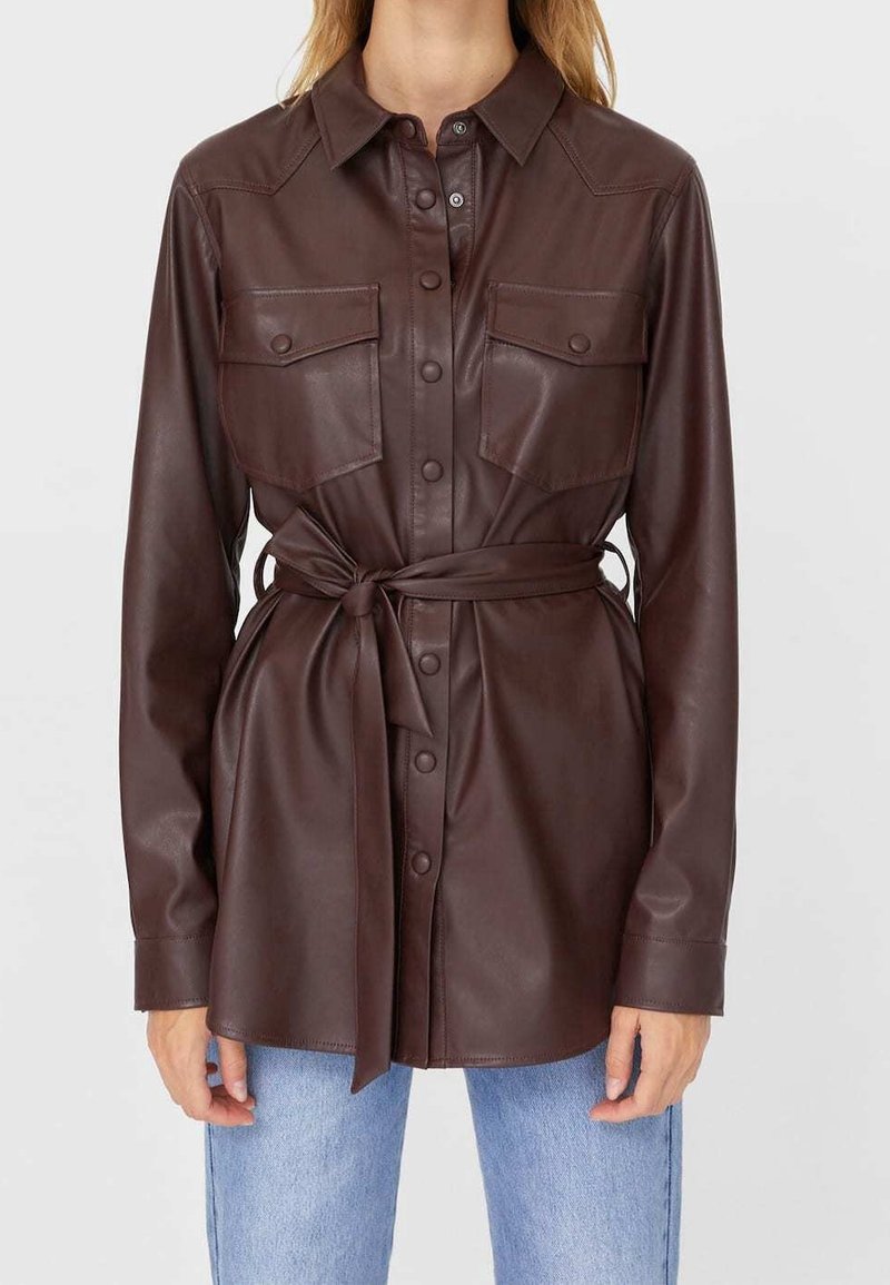 Brązowa skórzana koszula-kurtka z kołnierzykiem, zapinana na guziki z dwoma kieszeniami na piersi i paskiem wiązanym w talii.