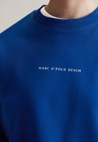 Blauwe sweatshirt met een geribbelde ronde hals. Voorzien van witte tekst "MARC O'POLO DENIM" op de voorkant. De stof lijkt glad en duurzaam.