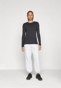 Frau, die in einem schwarzen Langarmshirt, hellgrauen Sweatpants mit elastischen Bündchen und schwarzen Sportschuhen vor einem schlichten weißen Hintergrund steht.