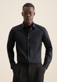 Camicia nera a maniche lunghe con bottoni, dalla texture liscia. Presenta un colletto classico e bottoni argentati. Indossata con pantaloni neri plissettati.