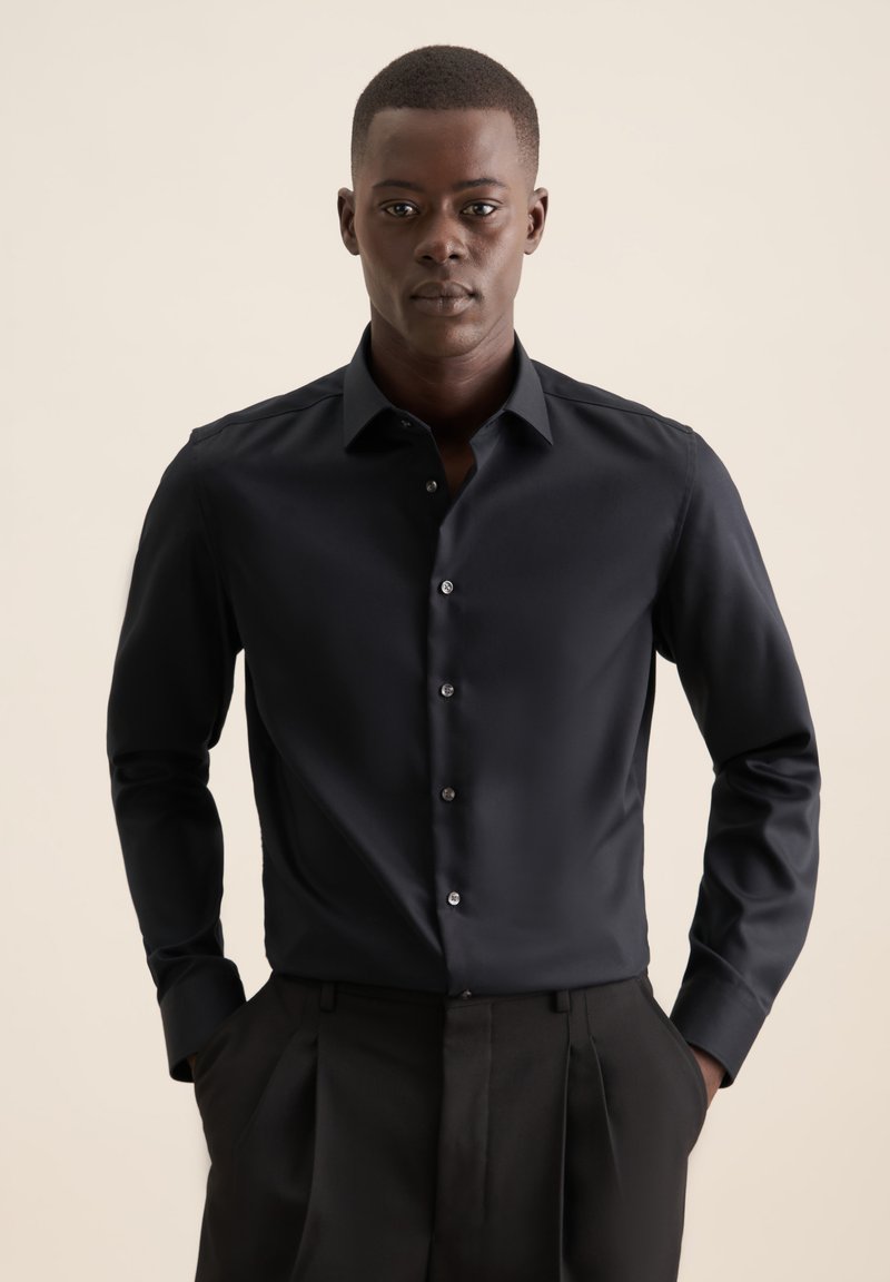 Camicia nera a maniche lunghe con bottoni, dalla texture liscia. Presenta un colletto classico e bottoni argentati. Indossata con pantaloni neri plissettati.