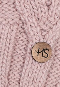 Knitted pink fabric with thick, textured yarn featuring a prominent circular metal tag embossed with "HS" in black lettering.
