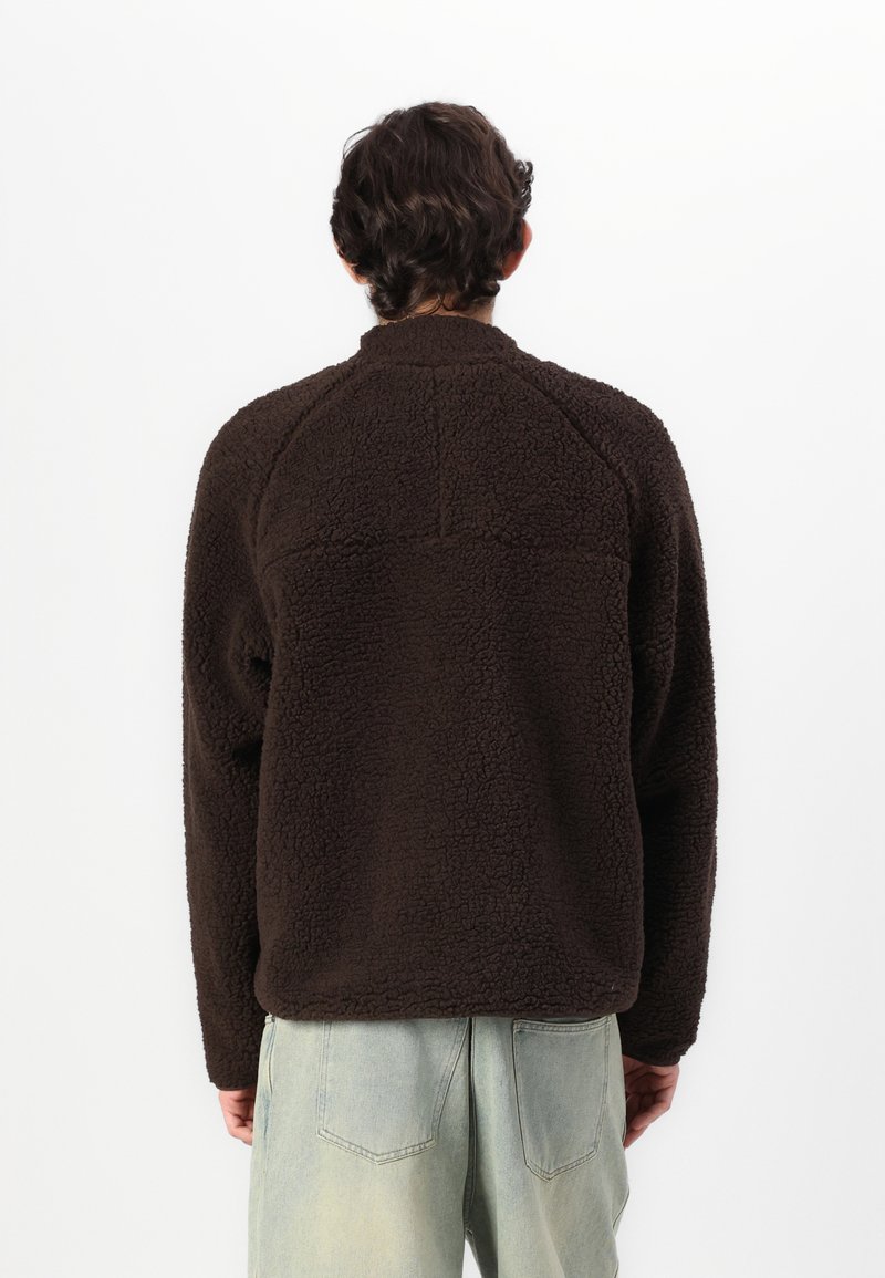 Brązowy polarowy sweter z teksturowanej powierzchni i wysokim kołnierzem, charakteryzujący się luźnym krojem i rękawami raglanowymi. Widok z tyłu.