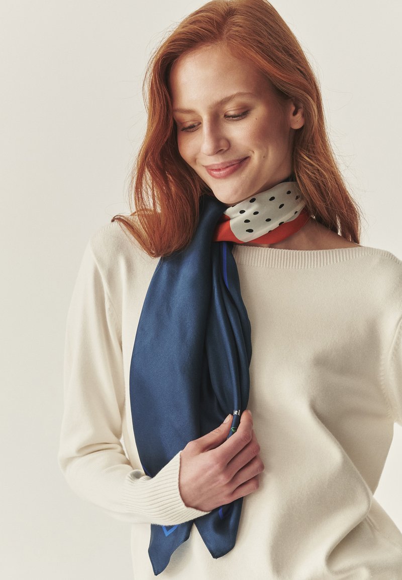 Sciarpa di seta blu navy con motivo a pois arancioni, elegantemente drappeggiata intorno al collo, abbinata a un maglione color crema.