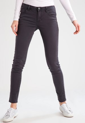 Person wearing slim-fit dark gray jeans and white sneakers, standing with legs apart against a plain light background.