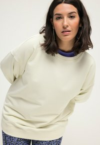 Sudadera de gran tamaño en verde claro con escote redondeado, mangas con puños y textura suave, superpuesta sobre una camiseta morada y leggings con estampado.