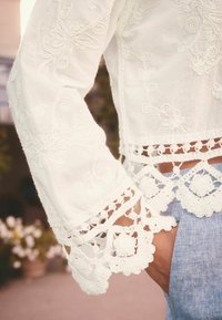 White embroidered blouse with long sleeves, featuring intricate floral patterns and lace accents at the cuffs and hem. Paired with denim.