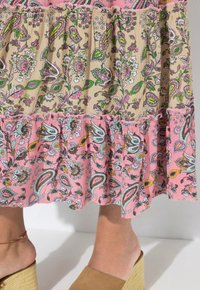 Robe à motifs floraux avec des volants superposés en rose et beige, ornée d'accents verts et bleus. Modèle portant des sandales à talon compensé marron clair.