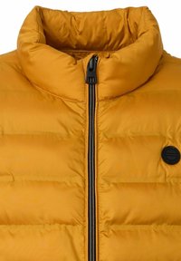 Gelbe Pufferjacke mit hohem Kragen, durchgehendem Reißverschluss und schwarzem Zubehör. Verfügt über eine gesteppten Textur und ein kreisförmiges schwarzes Logo auf der Brust.
