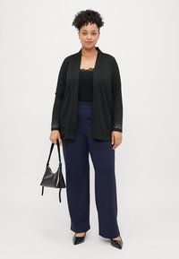 Μαύρος καρδιοκυματιστός με υφασμάτινα μανίκια, φορεμένος πάνω από ένα τοπ. Συνδυασμένος με ναυτικές παντελόνες ευρείας εφαρμογής και μαύρα παπούτσια με μυτερές μύτες. Κρατά μια μαύρη τσάντα.