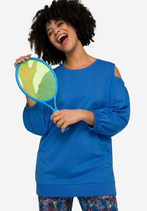 Felpa blu a maniche lunghe con design a spalle scoperte, abbinata a leggings con motivo vivace e a una racchetta blu e verde.