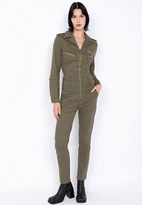 Combinaison vert olive avec fermeture éclair à l'avant, col et deux poches zippées. Elle présente une coupe ajustée et est associée à des bottines noires.