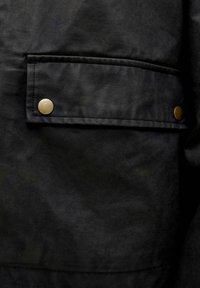 Czarna materiałowa kurtka z wyraźną kieszenią na piersi z mosiężnym zapięciem na zatrzask i gładkim, lekko teksturowanym wykończeniem.