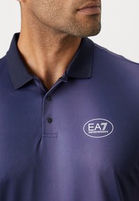 Marineblå polo skjorte laget av glatt stoff, med en to-knapps placket og en hvit brodert EA7 Emporio Armani-logo på brystet.